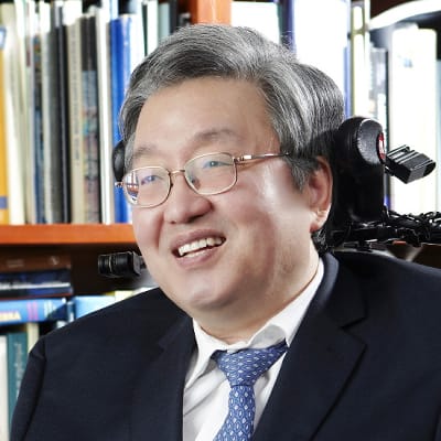 scientist with grey hair and glasses siting in a suit smiling 