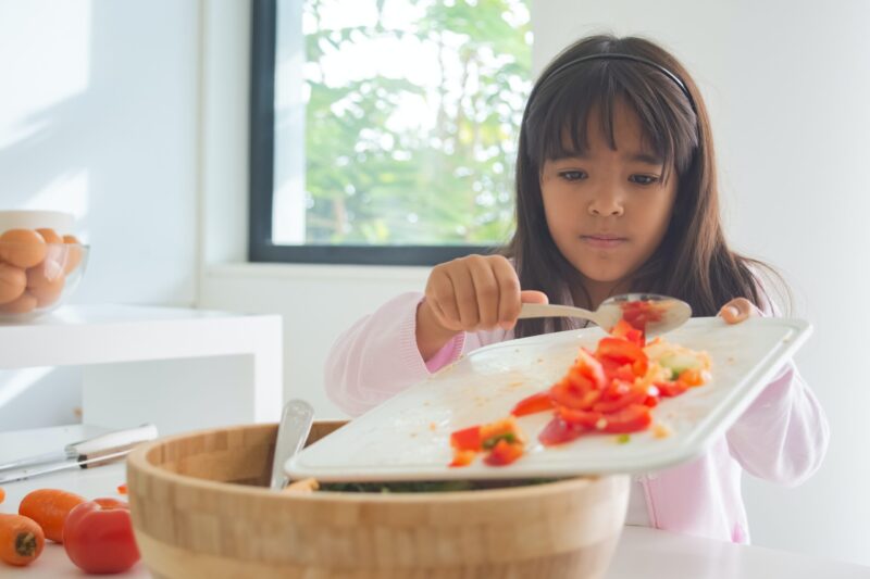 child scrapping cut up vegetables into a bowl