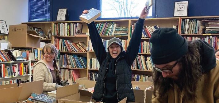 three patrons surrounded by books at the Great Book Grab, one woman is in the middle cheerful with her arms in the air