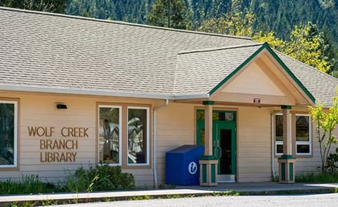 Small white building with a simple peak entry and the library logo on the wall with a forest in the background.